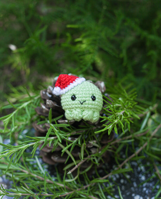 Crochet mini doll amigurumi styled as a festive Christmas ornament, hanging on a holiday-themed backdrop to demonstrate its use in seasonal decor